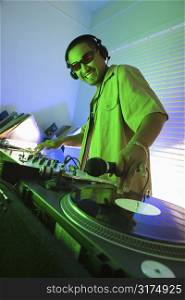 Asian young adult male DJ wearing sunglasses behind mixing equipment with hand on record turntable looking at viewer smiling.