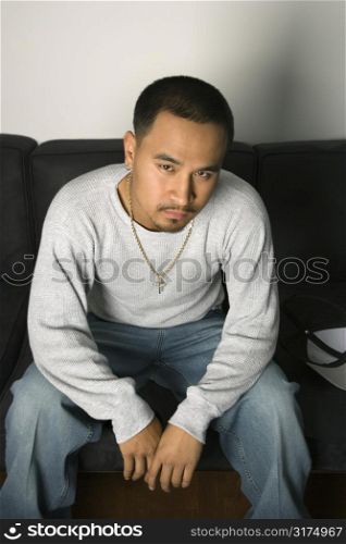 Asian young adult man sitting on sofa looking at viewer with critical expression.