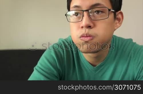 Asian young adult man watching TV and changing channel with remote control. The guy is bored and shakes head disappointed