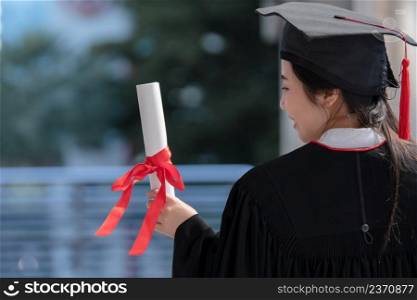 Asian young beautiful graduate female student with University degree turn back standing and holding diploma in hand and looking with proud face after graduation. Education concept.