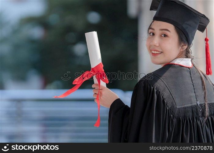 Asian young beautiful graduate female student with University degree turn back standing and holding diploma in hand and smiling with proud face after graduation. Education concept.