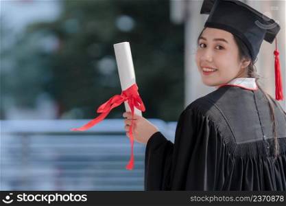 Asian young beautiful graduate female student with University degree turn back standing and holding diploma in hand and smiling with proud face after graduation. Education concept.