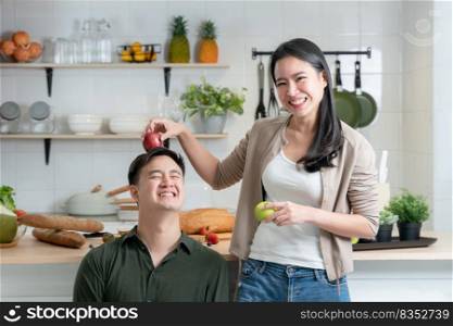 Asian young beautiful woman having fun with place a red apple on head of her handsome man in kitchen while cooking healthy food together. Cute teasing couple in modern kitchen concept