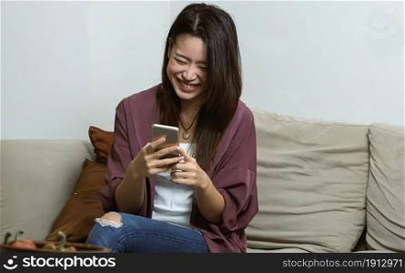Asian young beautiful woman smiling and chatting by using mobile phone while relaxing in living room at home. Lifestyle, Technology and New Normal Concept.