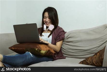 Asian young beautiful woman using laptop and headphone while relaxing in living room at home. Lifestyle, Technology and New Normal Concept.