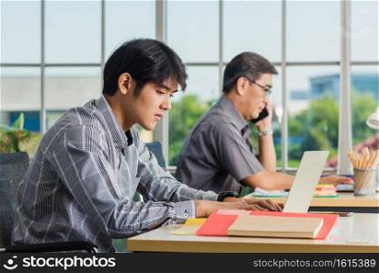 Asian young businessman sitting on desk office he looking at computer monitor during the working day, the confident young handsome man using laptop at workplace