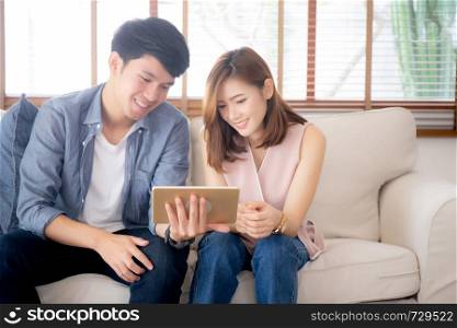 Asian young couple relax looking tablet entertainment on internet together on sofa at home, family leisure watching movie digital with comfortable, lifestyle and wellbeing concept.