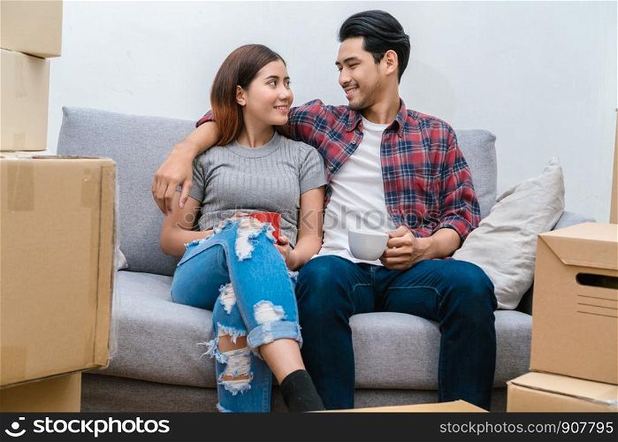 Asian young couple Sitting on the sofa and drinking for relax after success packing big cardboard box for moving in new house, Moving and House Hunting concept