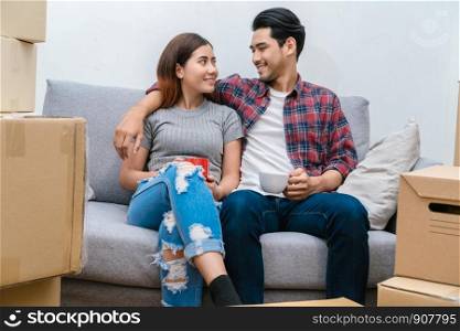 Asian young couple Sitting on the sofa and drinking for relax after success packing big cardboard box for moving in new house, Moving and House Hunting concept