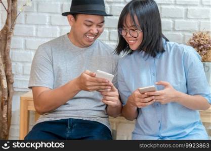 Asian Young Couple talking and using the smart mobile phone with happiness action in modern coffee shop or workplace or co-working space or modern office, lifestyle and leisure concept