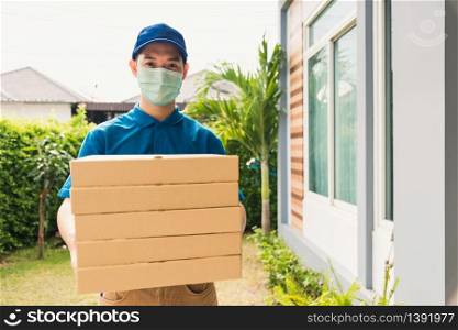Asian young delivery man courier sending and holding fast food pizza boxed in uniform he protective face mask service customer at home door, under curfew quarantine pandemic coronavirus COVID-19