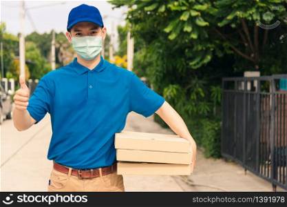 Asian young delivery man courier sending and holding fast food pizza boxed in uniform he protective face mask show thumb up for good sign, under curfew quarantine pandemic coronavirus COVID-19