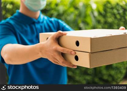 Asian young delivery man courier sending and holding fast food pizza boxed in uniform he protective face mask service customer at home door, under curfew quarantine pandemic coronavirus COVID-19