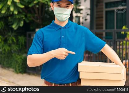 Asian young delivery man courier sending and holding fast food pizza boxed in uniform he protective face mask pointing finger to pizza box, under curfew quarantine pandemic coronavirus COVID-19