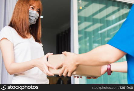 Asian young delivery man courier with package post box in uniform he protective face mask he making service woman customer receiving front house under curfew quarantine pandemic coronavirus COVID-19