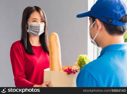 Asian young delivery man in uniform wear protective face mask he making grocery service giving fresh food to woman customer receiving front house under pandemic coronavirus, Back to new normal concept