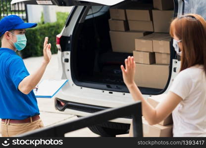 Asian young delivery man raises hand for bye after woman customer received boxes, both protective face mask under curfew quarantine pandemic coronavirus COVID-19 at front home door