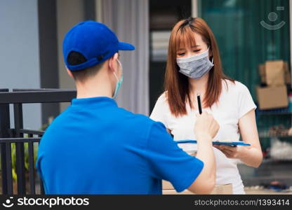 Asian young delivery man with package post boxed he protective face mask service woman customer using pen signature on paper book at front home, under curfew quarantine pandemic coronavirus COVID-19