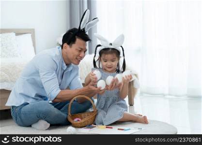 Asian young father and litt≤cute daughter wearing bunny ears and enjoy pa∫ing on eggs for Easter holidays whi≤sitting on floor to≥ther at home. Happy Easter Family traditions concept