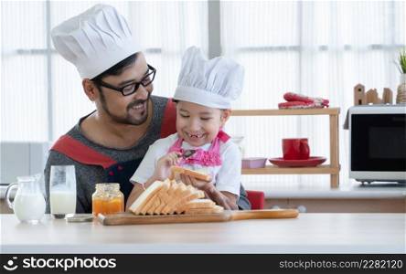 Asian young father with beard and little cute daughter with apron and chef hat smiling and preparing breakfast like spread jam on sliced bread together in kitchen at home. Lovely relationship concept