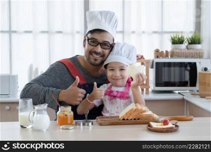 Asian young father with beard and little mixed race cute daughter with apron and chef hat smiling and thumb up holding heart shape bread while preparing breakfast together in kitchen at home