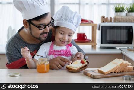 Asian young father with beard and little mixed race cute daughter with apron and chef hat smiling and preparing breakfast like spread jam on bread with strawberry together in kitchen at home