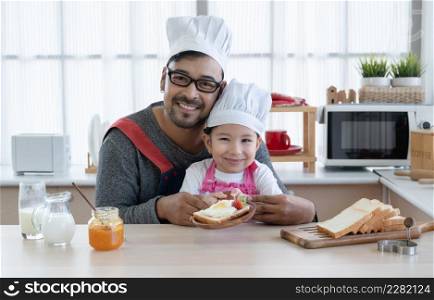 Asian young father with beard and little mixed race cute daughter with apron and chef hat smiling and showing spread jam on heart shape bread with strawberry together in kitchen at home