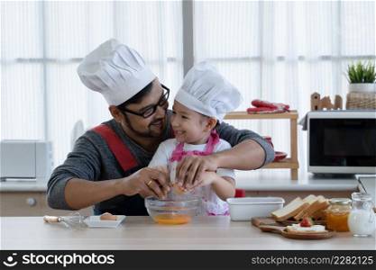 Asian young father with beard and little mixed race cute daughter with apron and chef hat smiling and cracking an egg in a bowl while preparing breakfast together in kitchen at home
