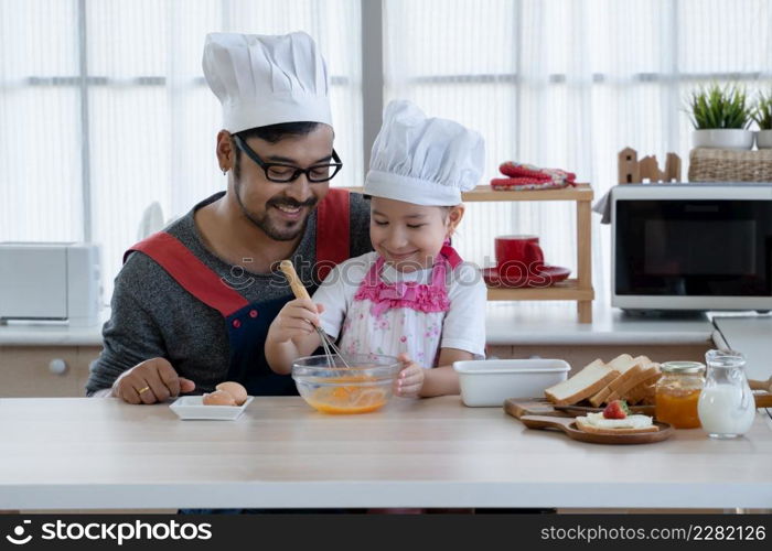 Asian young father with beard and little mixed race cute daughter with apron and chef hat smiling and holding whisk to mix eggs in a bowl while preparing breakfast together in kitchen at home