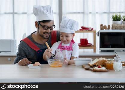 Asian young father with beard and little mixed race cute daughter with apron and chef hat smiling and holding whisk to mix eggs in a bowl while preparing breakfast together in kitchen at home