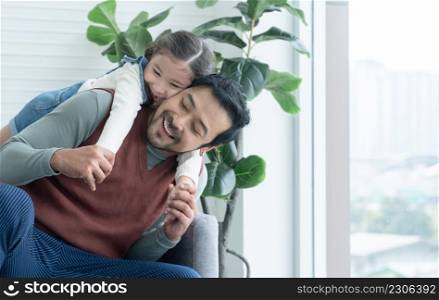 Asian young father with beard enjoy playing with little Caucasian kid girl in living room at home. Happy daughter hugging and riding on back of dad . Diverse family concept