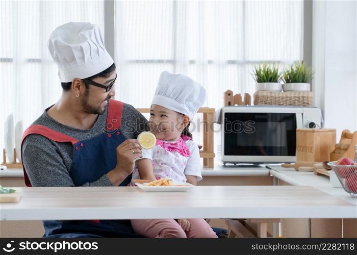 Asian young father with beard smiling and giving lemon slice to little cute mixed race daughter with apron and chef hat to try taste while enjoy eating spaghetti together in kitchen at home