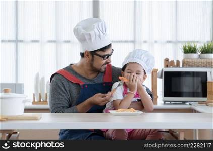 Asian young father with beard smiling and giving tomato slice to little cute daughter with apron and chef hat try taste but kid deny by covered her mouth with hands while eating spaghetti in kitchen