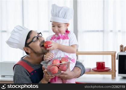 Asian young father with beard smiling and holding red apples basket in hands and little cute daughter with apron and chef hat giving dad an apple to eat in kitchen at home. Lovely relationship concept