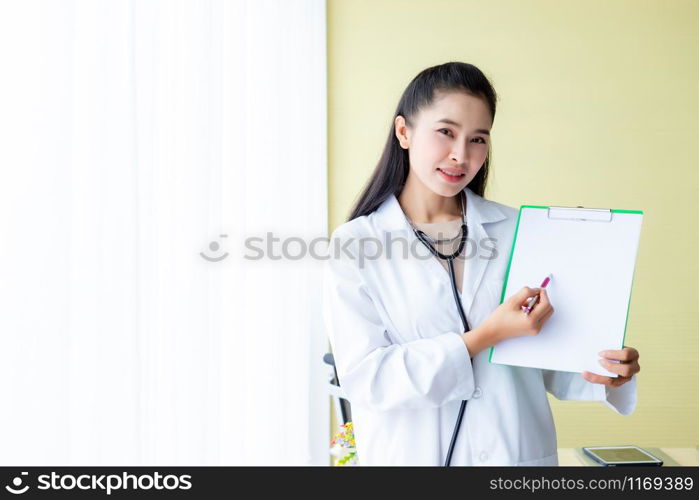 Asian young female doctor therapeutic advising with positive emotions hold empty clipboard and bed in hospital background,copy space