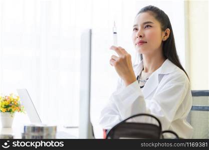 Asian young female doctor with stethoscope with holding a syringe and laptop computer,analogue pressure gauge on wooden table in Hospital background