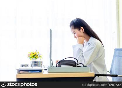 Asian young female doctor with stethoscope working with laptop computer and analogue pressure gauge on wooden table in Hospital background