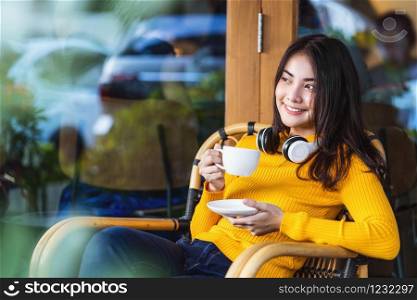 Asian young female holding a cup of coffee and sitting in modern coffee shop or coworking space beside window mirror, wearing Headphones for listening the music,hipster lifestyle and freelance concept
