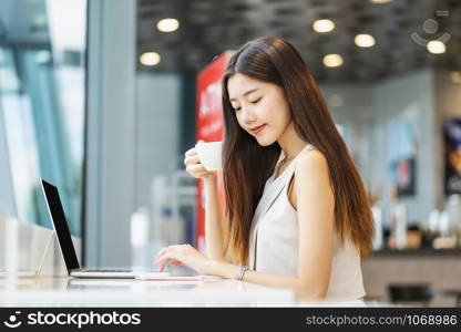 Asian young female holding a cup of coffee and using mobile phone with technology laptop in modern coffee shop or coworking space, technology smart mobile and hipster lifestyle, entrepreneur concept