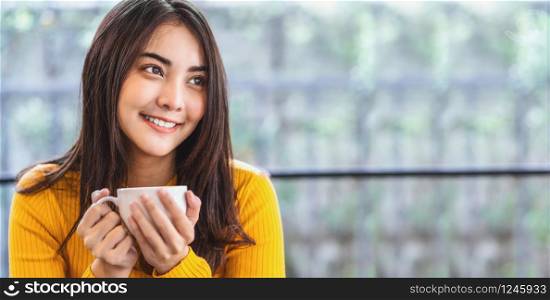Asian young female holding a cup of coffee in modern coffee shop or coworking space beside window mirror, Lifestyle and leisure with hobby and entrepreneur concept, banner or web cover scale
