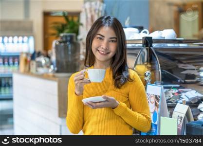 Asian young female holding a cup of coffee in modern coffee shop or coworking space beside coffee espresso machine, Lifestyle and leisure with hobby and entrepreneur concept