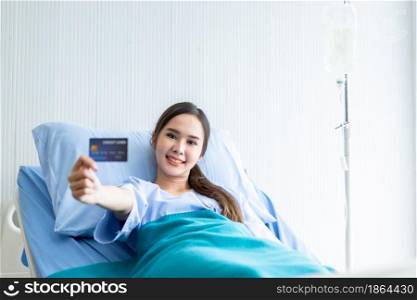 Asian young female patient smiley face on show holding a credit card lying on bed in the room hospital background,payment medical treatment concept