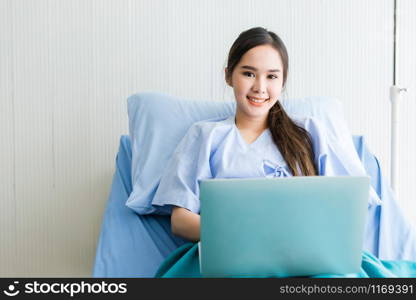 Asian young female patient with a smile working with laptop computer on bed in the room hospital background,concept of working business in a sick time