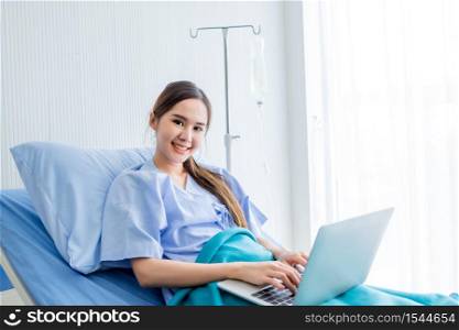 Asian young female patient with a smile working with laptop computer on bed in the room hospital background,concept of working business in a sick time