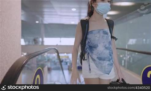 Asian Young female wear protective face mask walking out from escalator with backpack inside empty airport terminal, risk of infectious diseases at public places, new normal pandemic, travel insurance