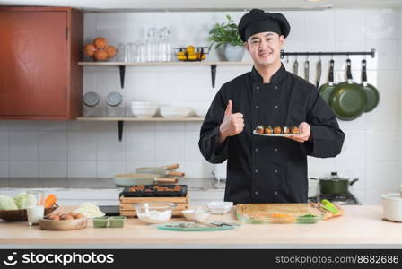 Asian young handsome chef man, in black uniform, smiling thumbs up and holding on japanese food called takoyaki in plate ready to serve from hot pan with ingredients on table at kitchen restaurant