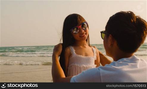 Asian young happy family enjoy vacation on beach in the evening. Dad help kid wears sunglasses while relax together near sea when sunset. Lifestyle travel holiday vacation summer trip concept.