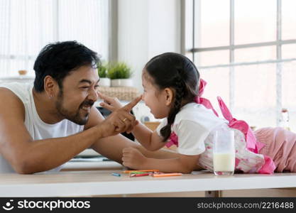 Asian young healthy dad with beard and little mixed race daughter smiling playing touching nose each other in kitchen at home with a glass of milk and color pencils on table