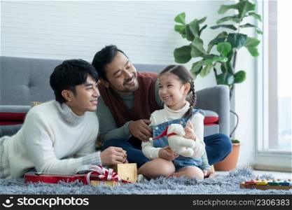 Asian young LGBTQ gay couple giving gift to little Caucasian adopted kid in living room at home. Kid smiling and delighted to receive doll as a present on birthday or special day. LGBT family concept