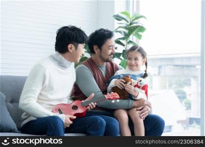 Asian young LGBTQ gay couple playing ukulele and singing with little Caucasian adopted kid in living room at home. Daughter holding dolls and sitting on dad lap on sofa. LGBT diverse family concept